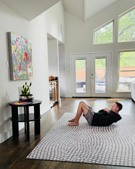Person doing sit-ups on the Circa Sandstone oversized exercise mat, a non-toxic high-density foam mat in a warm sandstone pebble pattern, during a home workout