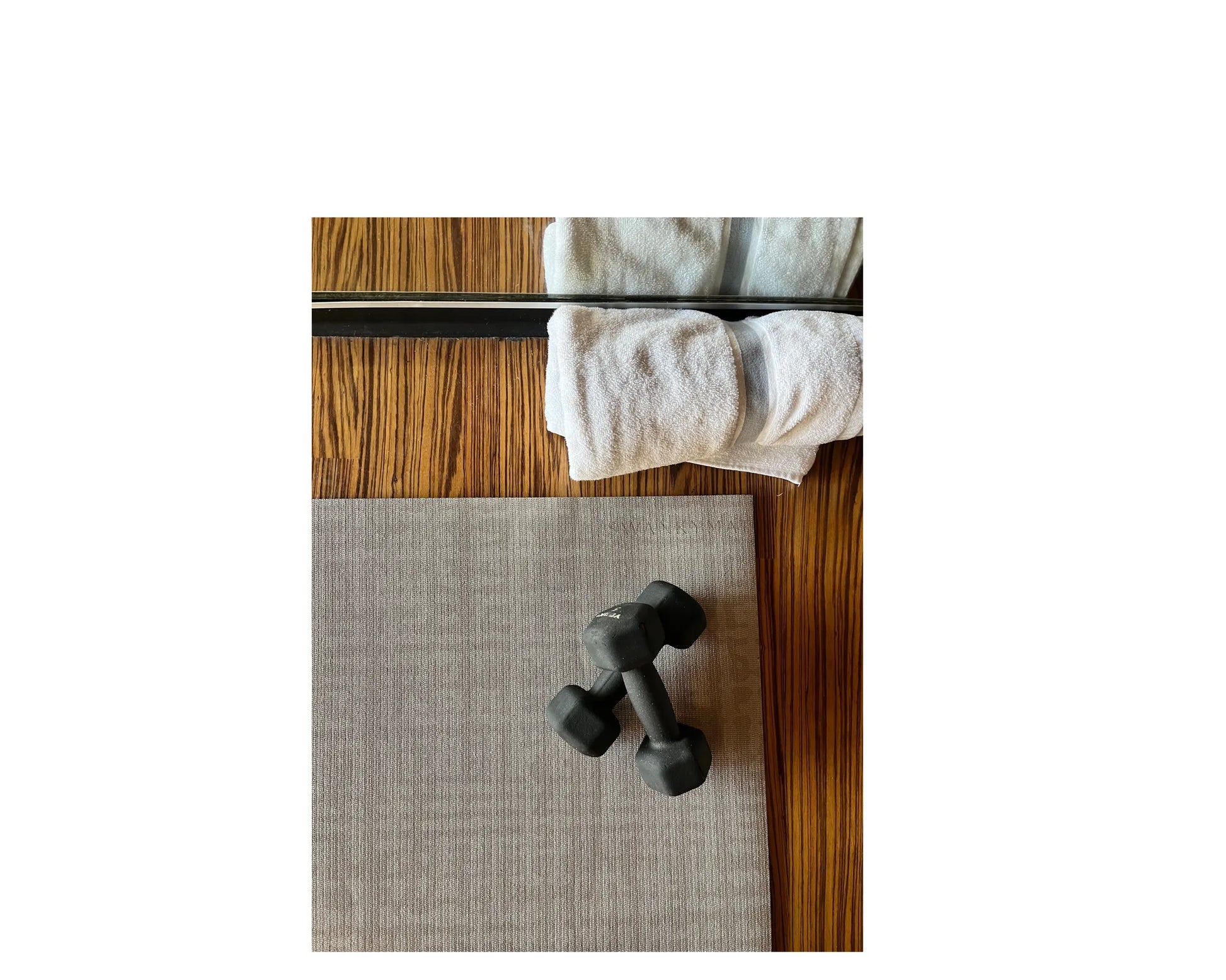 Black dumbbells and rolled white towels on the Swankymat Sloane Dust Studio Mat — a non-toxic, high-density foam yoga and pilates mat in a soft greige crosshatch, styled on a wood floor in a home studio.