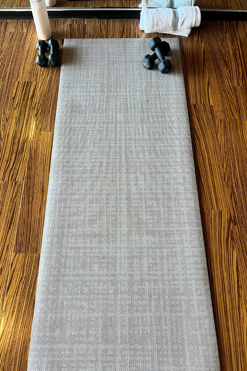 Swankymat Sloane Dust Studio Mat rolled out full-length on a wood floor with dumbbells at one end — a rollable, non-toxic high-density foam exercise mat for yoga, pilates, and HIIT in a neutral Dust crosshatch.